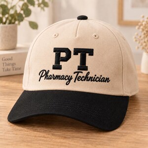 May include: A beige baseball cap with a black brim and a black button on top. The front features the embroidered black text PT in a block font, with the words Pharmacy Technician embroidered below in a black cursive script font.