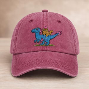 May include: A maroon baseball cap featuring an embroidered graphic of a blue dinosaur with pink accents. A brown cat wearing a small pink crown and black sunglasses is riding on the dinosaur's back. The design includes green grass below.