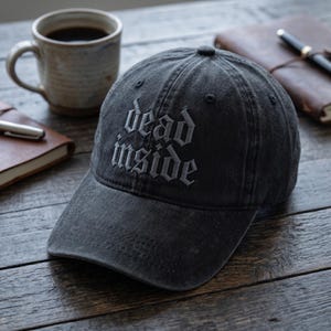 May include: A charcoal grey baseball cap with a distressed, washed-out finish. The front of the cap features the text "dead inside" embroidered in a gothic-style font. The cap has a curved brim, a button on top, and stitched eyelet details.