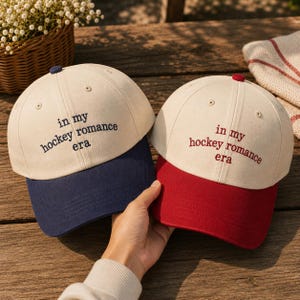 May include: Two baseball caps with off-white crowns and embroidered text reading "in my hockey romance era". One cap features a dark blue brim and top button, while the other cap features a dark red brim and top button, both with matching embroidery.