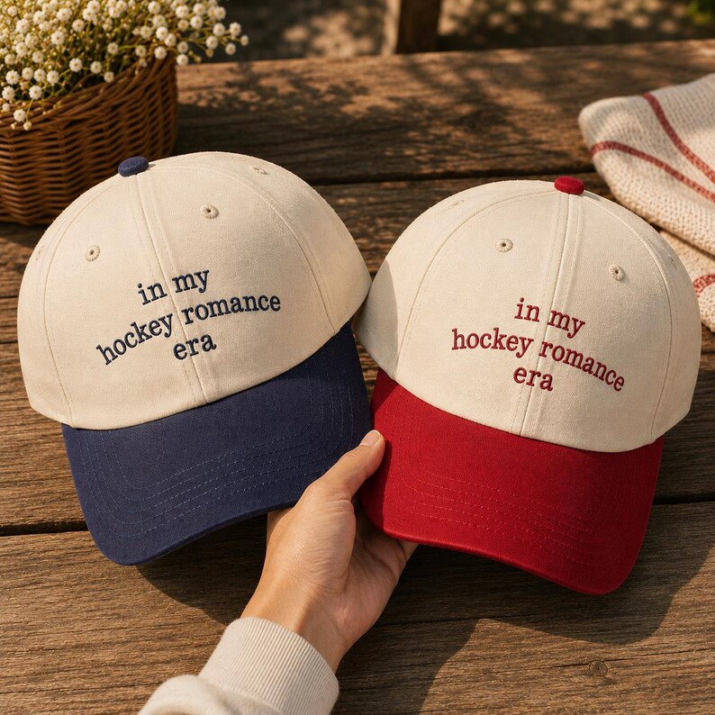 May include: Two baseball caps with off-white crowns and embroidered text reading "in my hockey romance era". One cap features a dark blue brim and top button, while the other cap features a dark red brim and top button, both with matching embroidery.
