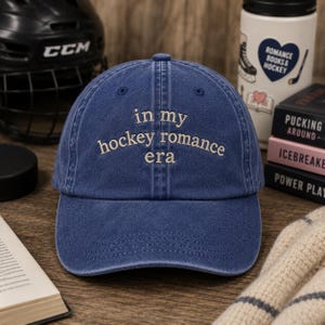 May include: A blue baseball cap with a distressed finish featuring the embroidered text "in my hockey romance era" in a light beige color. The cap has a structured front panel, a curved brim, and a button on the top center of the crown.