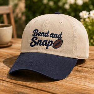 May include: A beige baseball cap with a dark blue brim and embroidered dark blue text that reads Bend and Snap, accompanied by a small brown football graphic. The cap features a structured crown with a button on top and stitched eyelets.