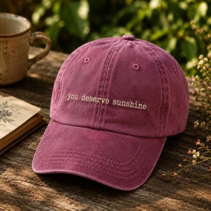 May include: A deep magenta baseball cap featuring the embroidered text you deserve sunshine in a light cream colour. The cap has a structured crown with ventilation eyelets and a curved brim, showcasing a washed fabric texture throughout.