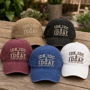 May include: Five baseball caps in tan, black, maroon, blue, and white. Each cap features embroidered text that reads IDK, IDC & IDGAF followed by the phrase LIVING MY BEST LIFE. The caps have a curved brim and a structured crown with eyelets.