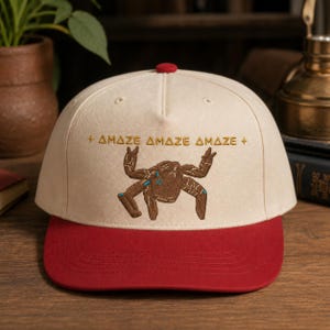 May include: A cream-colored baseball cap with a red brim and a red button on top. The front features an embroidered brown crab-like creature with blue accents, flanked by small star symbols and the repeated text "AMAZE AMAZE AMAZE" in gold.