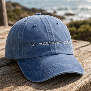May include: A blue denim baseball cap with the embroidered text "to whatever end" in white. The cap has a curved peak and a button on top. The background shows a blurred view of the sea and foliage.