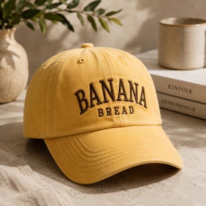 May include: Mustard yellow baseball cap with the embroidered text "BANANA BREAD" in brown. The cap has a curved peak and a button on top. A casual hat.