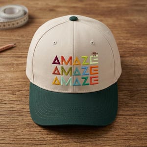 May include: A beige baseball cap with a dark green brim and a dark green button on top. The front features the word AMAZE embroidered in three rows using various colors including red, orange, yellow, light green, blue, and teal, with a small brown figure.