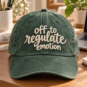 May include: A dark green baseball cap with a washed, distressed texture. The front features embroidered cream-coloured text that reads "off to regulate emotion" in a mix of block and cursive lettering. The cap has a structured crown and a curved brim.