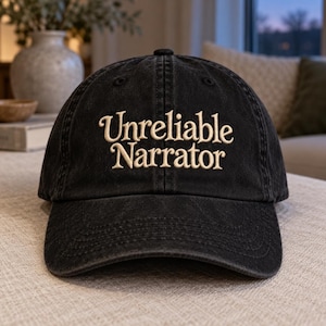 May include: A black baseball cap with a washed, vintage-style finish. The front features the text "Unreliable Narrator" embroidered in a light beige, serif font. The cap has a structured crown, a curved brim, and visible stitching along the panels.