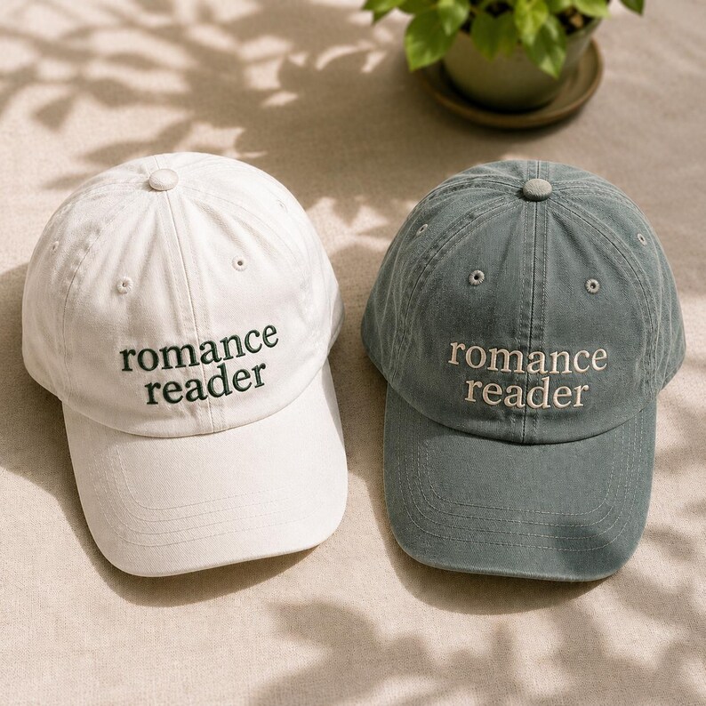 May include: Two baseball caps, one white and one muted blue-green, each featuring the embroidered text "romance reader" in a contrasting color on the front panel. Both caps have a structured crown, a curved brim, and a button at the top center.