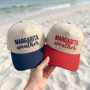 Peut inclure: Deux casquettes de baseball beiges avec du texte brodé. L'une présente le texte bleu foncé MARGARITA weather et une visière bleu foncé. L'autre présente le texte rouge MARGARITA weather et une visière rouge. Les deux ont une calotte structurée.