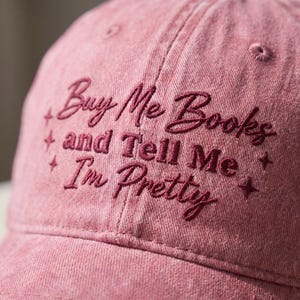 May include: A dusty rose baseball cap featuring dark red embroidered text that reads Buy Me Books and Tell Me I'm Pretty, accented by small embroidered star shapes on both sides of the lettering, with visible stitching and eyelet details.