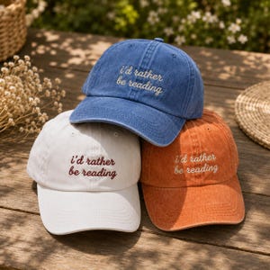 Peut inclure: Trois casquettes de baseball en bleu, blanc et orange, chacune portant le texte brodé "i'd rather be reading" sur le panneau avant. La casquette bleue a du texte blanc, la blanche a du texte rouge foncé et l'orange a du texte blanc.