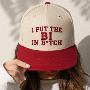 Peut inclure: Une casquette de baseball beige avec une visière plate rouge et un texte brodé en rouge sur le devant qui indique I PUT THE BI IN B*TCH. La casquette présente une calotte structurée avec un petit bouton rouge sur le dessus et des coutures rouges.