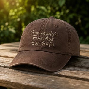 Peut inclure: Une casquette de baseball marron avec une visière incurvée et une construction à six panneaux. L'avant de la casquette présente un texte brodé de couleur beige clair qui indique, Somebody's Fine Ass Ex-Wife, dans une police cursive.
