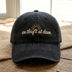 Peut inclure: Une casquette de baseball noir anthracite avec une finition délavée. L'avant présente une broderie dorée d'une icône de soleil levant au-dessus du texte "we thrift at dawn" écrit en écriture cursive. La casquette a une visière courbée.