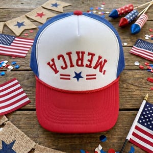 Könnte beinhalten: Eine rot-weiß-blaue Trucker-Cap mit weißem Vorderteil, auf dem der rote Schriftzug 'MERICA' über einem blauen Stern und zwei roten horizontalen Linien gestickt ist. Die Kappe hat einen roten Schirm, blaue Netzeinsätze und eine rote Kordel.