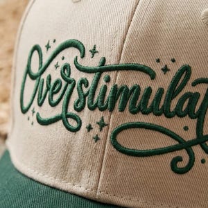 Peut inclure: Une casquette de baseball beige avec une inscription brodée en cursive vert foncé qui lit Overstimulated, agrémentée de petites étoiles brodées. La casquette présente une calotte structurée avec des coutures visibles et une visière vert foncé.