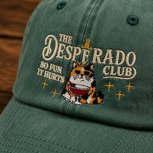 May include: A dark green baseball cap featuring embroidered text that reads THE DESPERADO CLUB SO FUN IT HURTS. The design includes a calico cat wearing sunglasses and holding a red bowl, accented by small yellow star shapes and a tiny sword icon.