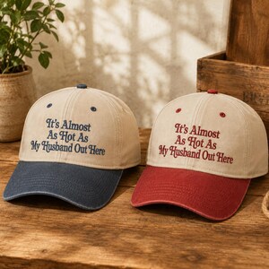 May include: Two baseball caps with beige crowns and embroidered text that reads It's Almost As Hot As My Husband Out Here. One cap features a navy blue brim and navy blue text, while the other has a red brim and red text. Both have top buttons.