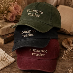 May include: A stack of three baseball caps in dark green, black, and maroon. Each cap features the text "romance reader" embroidered in a light beige thread on the front panel. The caps have a soft, washed fabric texture and a curved brim.