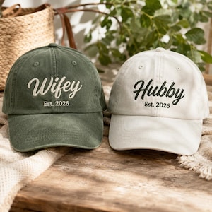 May include: A dark olive green baseball cap with white embroidered text reading Wifey Est. 2026 and a cream baseball cap with dark green embroidered text reading Hubby Est. 2026. Both caps feature a curved brim and a structured six-panel design.
