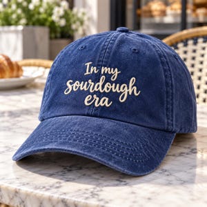 May include: A navy blue baseball cap with a distressed, washed-out finish. The front features the embroidered text "In my sourdough era" in a cream-coloured, cursive script font. The cap has a curved brim and a structured six-panel crown design.