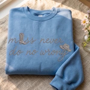Op de afbeelding: Een lichtblauwe sweatshirt met de geborduurde tekst "Miss never do no wrong". Het ontwerp bevat een cowboylaars en een cowboyhoed. De sweatshirt is op een neutraal gekleurd oppervlak gevouwen.