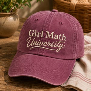 May include: A maroon baseball cap with a distressed, washed-out finish. The front features embroidered text in a cream-colored thread that reads Girl Math University, with the word University written in a cursive script font.