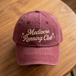 May include: A maroon baseball cap with a washed-out texture featuring the embroidered text "Mediocre Running Club" in a cream-colored script font, accented by small four-pointed star symbols on both sides of the lettering.