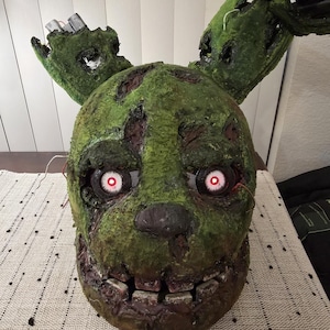 May include: A weathered, green, rabbit-like mask with a distressed, textured surface. It features two long ears, one partially broken, and glowing red circular eyes. The mouth area shows square teeth, and exposed wires are visible near the ears.