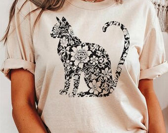 May include: Beige t-shirt featuring a black floral cat silhouette. The cat design is filled with intricate floral patterns. The shirt has rolled-up sleeves and a crew neck. The overall style is casual and comfortable.