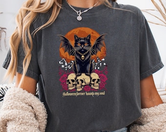 May include: Dark gray t-shirt featuring a black cat with bat wings, a skull necklace, and a full moon graphic. The cat sits atop three skulls surrounded by roses. Text reads "Halloween forever haunts my soul."