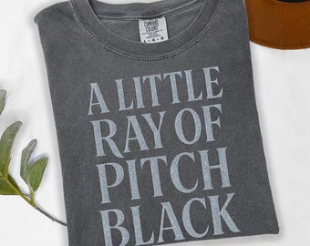 May include: Dark gray Comfort Colors t-shirt with the text "A LITTLE RAY OF PITCH BLACK" in silver lettering. The shirt is folded and has a crew neck. A brown hat is partially visible in the background.