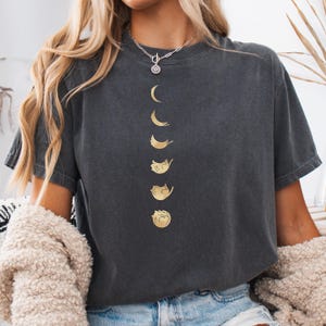 May include: Dark gray t-shirt featuring a gold design of a cat transforming into different moon phases. The shirt has a relaxed fit and short sleeves. The design is centered on the front of the shirt.