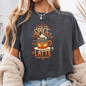 May include: Dark gray t-shirt featuring a fall-themed graphic. The design includes a pumpkin spice latte with the words "PUMPKABOO SPICE LATTE" in a decorative font. The graphic is in shades of orange, brown, and cream.