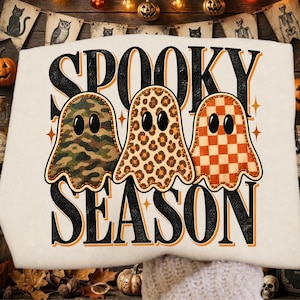 May include: A cream-colored sweatshirt featuring the black text SPOOKY SEASON and three ghosts. The first ghost has a green camouflage pattern, the second has a brown leopard print, and the third has an orange and white checkered pattern.