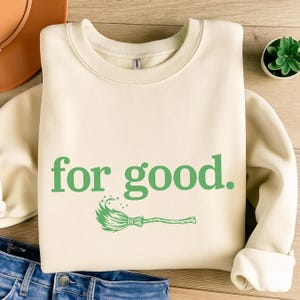 May include: Cream-colored sweatshirt with the phrase "for good." in green, along with a green witch's broom graphic. The sweatshirt is folded on a wooden surface, with a hat and jeans visible.