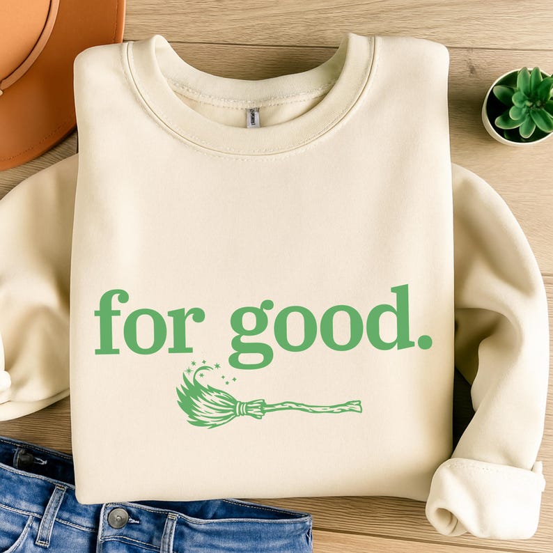 May include: Cream-colored sweatshirt with the phrase "for good." in green, along with a green witch's broom graphic. The sweatshirt is folded on a wooden surface, with a hat and jeans visible.