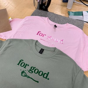 May include: Two t-shirts, one pink and one sage green, are displayed. The pink shirt has the words "for good" in a matching pink font, with a small design. The green shirt has the same text in green, with a broom graphic.