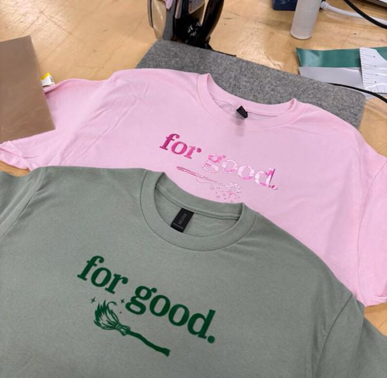 May include: Two t-shirts, one pink and one sage green, are displayed. The pink shirt has the words "for good" in a matching pink font, with a small design. The green shirt has the same text in green, with a broom graphic.