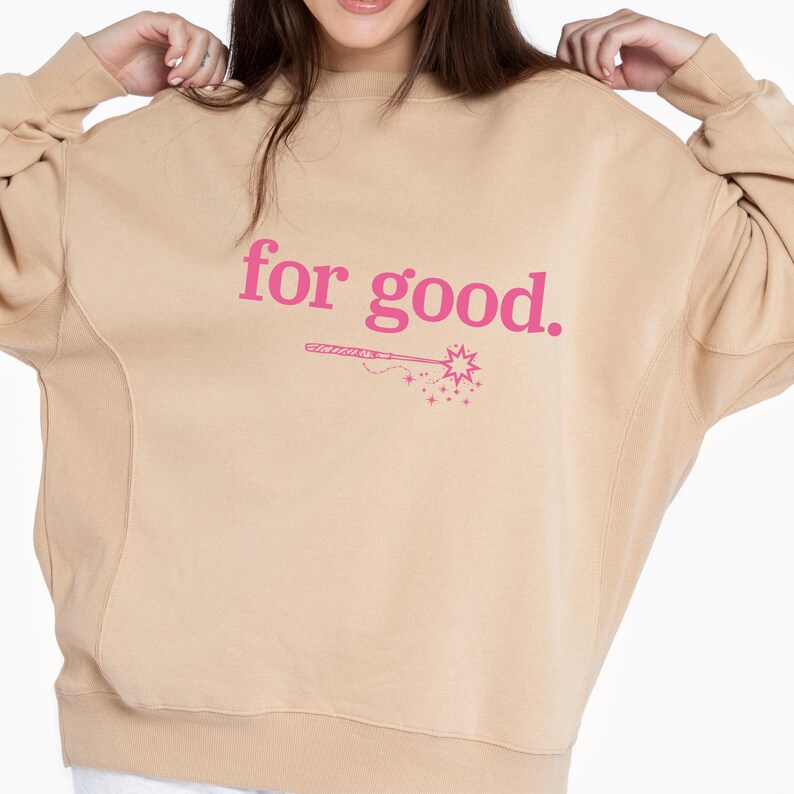 May include: A tan-colored sweatshirt with the phrase "for good." printed in pink, along with a pink illustration of a wand and stars. The sweatshirt has a crew neck and long sleeves.
