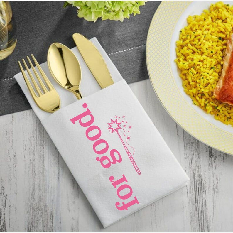 May include: A white napkin with the text "for good" in pink, along with a pink illustration of a magic wand. The napkin holds a gold fork, spoon, and knife. A plate of yellow rice and a piece of salmon is in the background.
