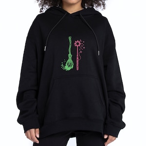 Op de afbeelding: Zwarte hoodie met een groene bezem en een roze toverstaf. De bezem staat links en de toverstaf rechts. Het ontwerp is gecentreerd op de voorkant van de hoodie.