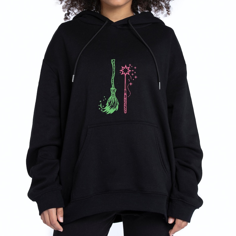 Op de afbeelding: Zwarte hoodie met een groene bezem en een roze toverstaf. De bezem staat links en de toverstaf rechts. Het ontwerp is gecentreerd op de voorkant van de hoodie.