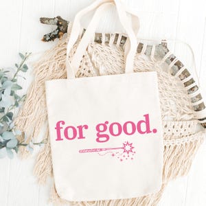 May include: Cream-colored tote bag with the phrase "for good." in bright pink lettering. Below the text is a pink illustration of a magic wand with stars. The bag has two cream-colored handles.