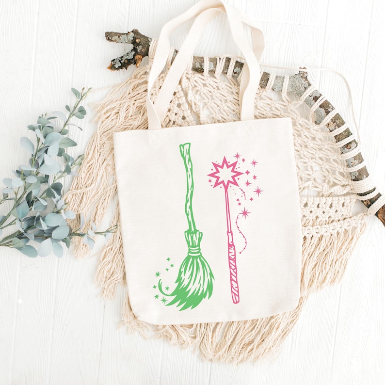 Op de afbeelding: Cr&egrave;mekleurige draagtas met een groen bezem- en een roze toverstafontwerp. De tas heeft lange, natuurlijk gekleurde handvatten. De achtergrond is wit met decoratieve elementen, waaronder macram&eacute; en bladeren.