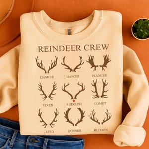 Puede incluir: Una sudadera beige con el texto "REINDEER CREW" e ilustraciones de cornamentas de reno. Las cornamentas están etiquetadas con nombres como "Dasher", "Rudolph" y "Blitzen". La sudadera está doblada sobre una superficie naranja.